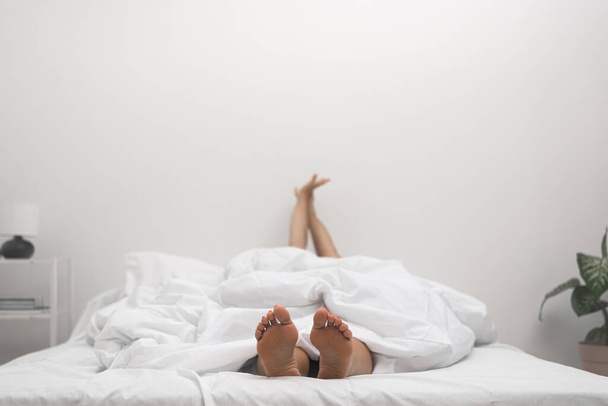 Жінка з голими ногами викладена з-під прикриття лежить на зручному ліжку в світлих тонах, що страждають від безсоння в освітленій спальні від хвороб житла з відсутністю сну в дорослому віці - Фото, зображення