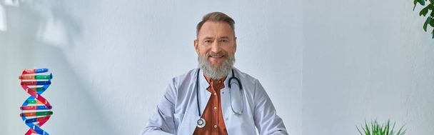 alegre médico barbudo gris sentado en la mesa mirando a la cámara en el fondo azulado, pancarta - Foto, Imagen