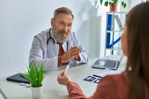 серйозний сірий бородатий лікар, уважно дивлячись на пацієнта жінки, що показує тест на вагітність, концепція IVf - Фото, зображення