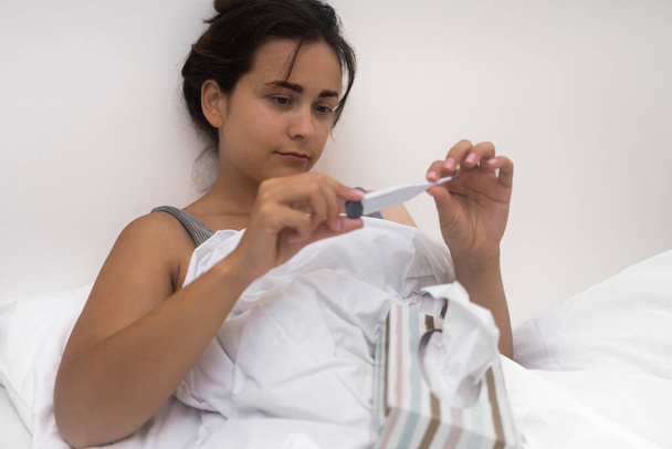 Νεαρή γυναίκα κοιτάζει το ηλεκτρονικό θερμόμετρο άρρωστο θηλυκό μετρά τη θερμοκρασία του σώματος κάθεται στο κρεβάτι με κουτί των ιστών του προσώπου έλεγχο για τα συμπτώματα της εποχιακής ασθένειας - Φωτογραφία, εικόνα