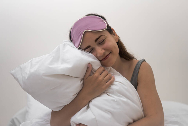Mulher jovem atraente sorri abraçando travesseiro branco fêmea feliz se prepara para a cama desfrutando de descanso em casa pessoa alegre com máscara de dormir senta-se na cama no quarto - Foto, Imagem
