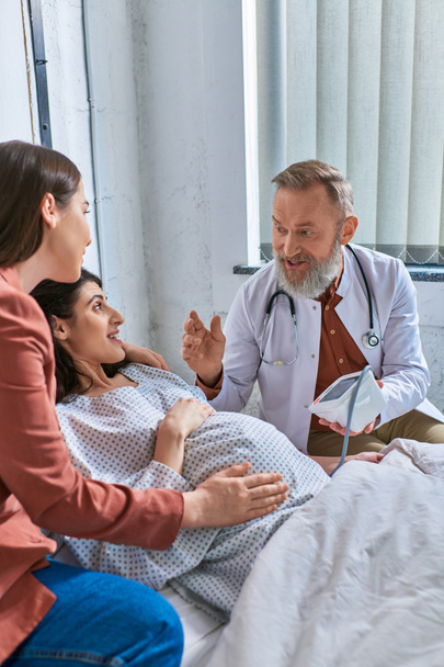 grijze bebaarde arts legt iets uit aan zijn zwangere patiënt meten druk, ivf concept - Foto, afbeelding