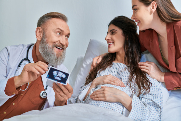smajlík šedý vousatý lékař ukazující ultrazvuk objímající pár Igbt při pohledu na ně, ivf koncept - Fotografie, Obrázek