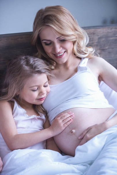 Schattig klein meisje is haar mooie zwangere Mama's buik aan te raken en glimlachen terwijl het besteden van tijd samen in bed - Foto, afbeelding
