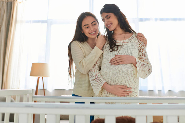 счастливая лесбийская пара тепло обнимается с рукой на беременном животе, стоя рядом с кроваткой, ivf концепция - Фото, изображение