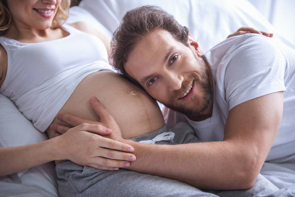 Krásná těhotná žena a její hezký manžel tráví čas spolu v posteli. Člověk poslouchá na miminko v břiše - Fotografie, Obrázek