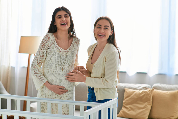 joyous lgbt couple standing next to crib smiling at camera with hands on pregnant belly, ivf concept - 写真・画像