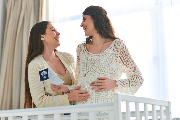 šťastný lesbický pár v blízkosti kolébky drží ultrazvuk a usmívá se upřímně na sebe, ivf koncept - Fotografie, Obrázek