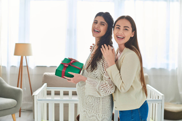 cheerful lesbian couple standing next to crib with gift in hands and smiling at camera, ivf concept - Photo, Image