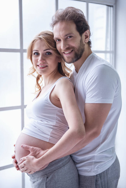 Hermosa mujer embarazada y su marido guapo están abrazando la barriga, mirando a la cámara y sonriendo
 - Foto, imagen