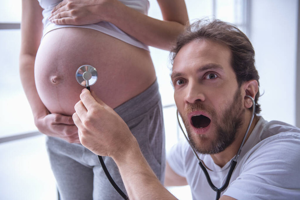 Handsome man is listening to his beautiful pregnant wife's tummy, using a stethoscope and showing shock - Photo, Image