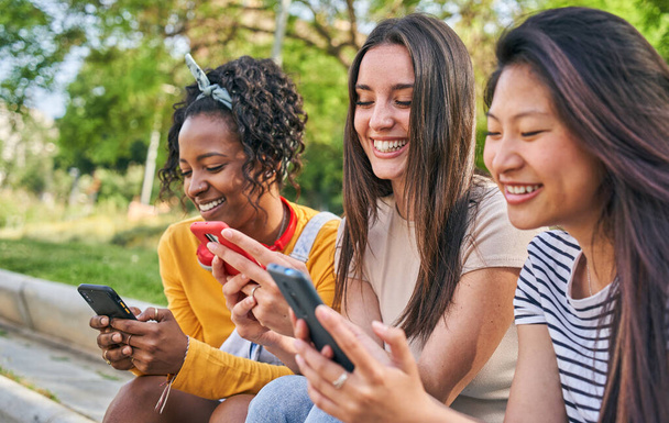 Three smiling young women using cell phone outdoors. Group of happy friends checking fun social networks on mobile. Cheerful classmates gossip on dating apps together. People and new technologies. - Photo, Image