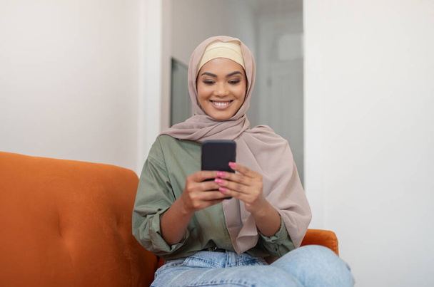 Aplicación móvil. Mujer negra feliz en Hijab usando el teléfono, mensajes de texto y chat en las redes sociales, sentado en el sofá en casa. Feliz dama islámica disfrutando del uso de Gadget navegar por Internet en el nuevo teléfono inteligente - Foto, imagen