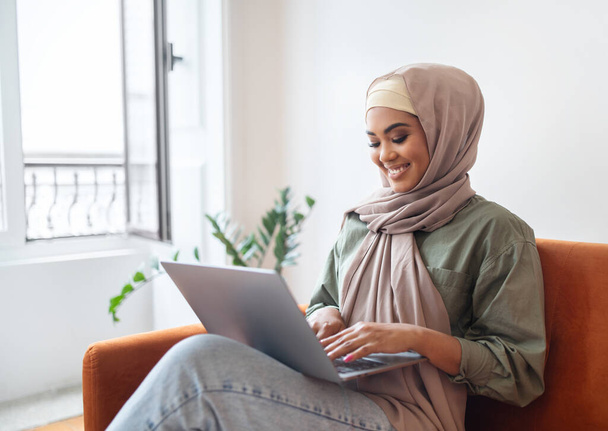 Carrera independiente. Mujer joven feliz en Hijab trabajando en línea en el ordenador portátil, escribiendo en la computadora, haciendo videollamada y aprendiendo sentado en el sofá en casa. Freelancer Websurf en la sala de estar - Foto, Imagen