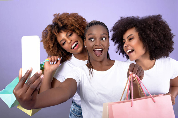 Shopping-Spaß. Drei aufgeregte junge schwarze Damen machen lustige Selfies per Handy, lächeln und halten Einkaufstaschen über lila Studiohintergrund. Gruppe weiblicher Käufer, tolles Vertriebskonzept - Foto, Bild