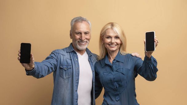 Cool online προσφορά για ηλικιωμένους. Χαρούμενος ηλικιωμένος άνδρας και γυναίκα ζευγάρι φίλους αγκαλιάζει και δείχνει τηλέφωνα με mockup λευκή μαύρη οθόνη, απομονώνονται σε μπεζ φόντο, χρονολόγηση mobile app - Φωτογραφία, εικόνα