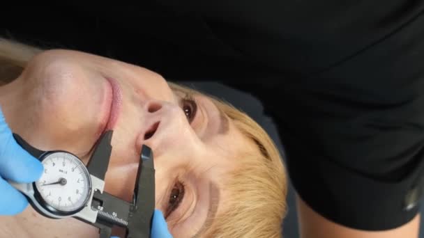 Lekarz przygotowuje starszą kobietę do operacji plastycznej twarzy. Perforacja na twarzy przed operacją plastyczną. Koncepcja chirurgii plastycznej twarzy. Pionowe wideo - Materiał filmowy, wideo