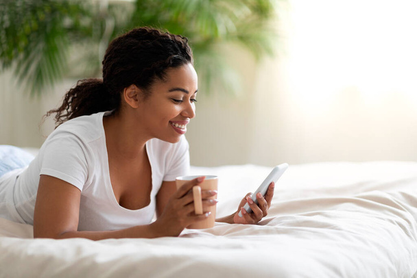 ナイスアプリ。 スマートフォンを使用して笑顔の若い黒人女性とベッドでコーヒーを飲む,ベッドルームで携帯電話でリラックスして幸せなアフリカ系アメリカ人女性,新しいアプリケーションをブラウズ,コピースペース - 写真・画像