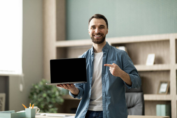 ハッピー ヤング ビジネスマン ポイント フィンガー アット ブランク ラップトップ スクリーン オフィスの近くに立っている間,スマイリング 男性起業家 ウェブサイト広告のための無料のコピー スペースを実証します., モックアップ - 写真・画像