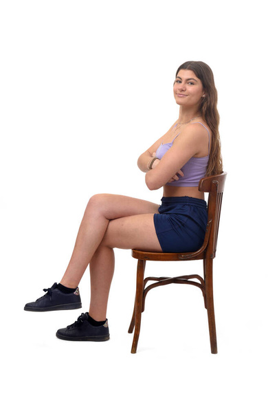 boční pohled na mladou dívku sedící na židli při pohledu na kameru a zkřížené nohy a paže zkřížené na bílém pozadí - Fotografie, Obrázek