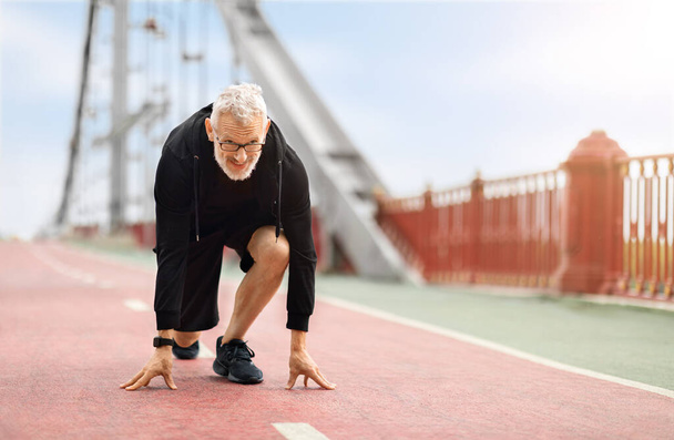 Běh, fitness, sport, outdoorové cvičení koncepce. Motivovaný atletický starší sportovec běžící po můstku, stojící na nízkém startu, dívající se dopředu, kopírující prostor. Zdravý lfestloue pro důchodce - Fotografie, Obrázek
