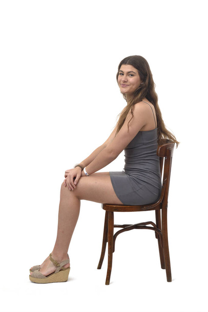 vista lateral de la joven sentada en la silla feliz y mirando a la cámara en el fondo blanco - Foto, Imagen