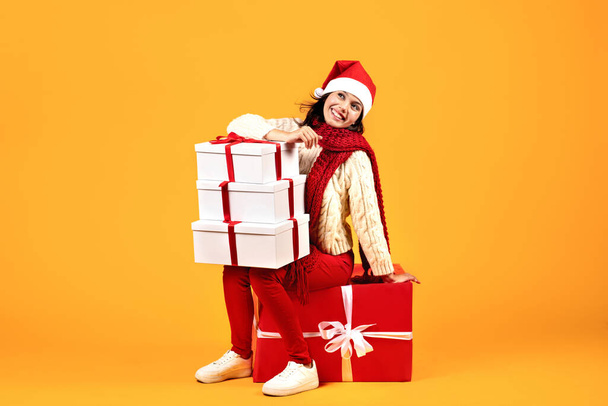 Mutlu Milenyum Kadını pozları, bir sürü Noel hediyesi kutusu, kırmızı Noel Baba şapkası takıyor ve sarı stüdyo arka planının üzerinde büyük bir hediye kutusunda oturuyor. Kış Tatil Satış ve Hediyeler Teslimatı - Fotoğraf, Görsel