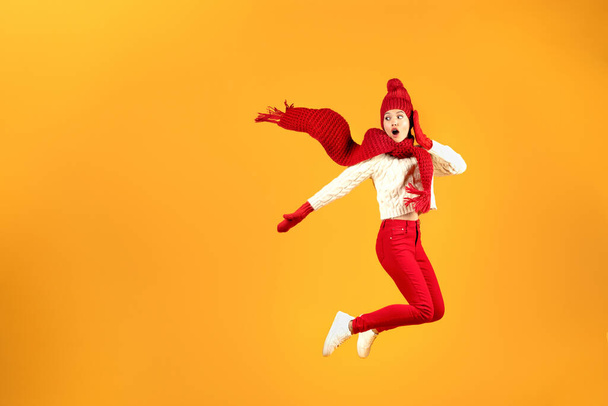 Great Winter Offer. Excited Young Woman In Red Knitted Hat And Scarf Jumping On Yellow Studio Background, Looking Aside At Empty Space, Posing In Mid Air. Christmas Season Sales Advertisement - Photo, Image