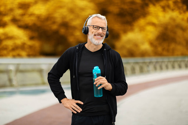 Retrato de deportista senior guapo alegre en ropa deportiva negra de pie en el parque de otoño, usando auriculares inalámbricos, sosteniendo la botella, agua potable durante el entrenamiento al aire libre, espacio para copiar - Foto, imagen