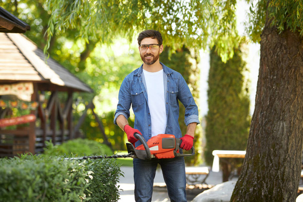 Happy male gardener in glasses posing, while holding hedge trimmer in park. Portrait of bearded man with electric bush cutter looking at camera, while working outdoors. Concept of seasonal work. - 写真・画像