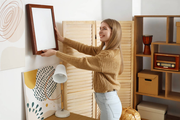jonge vrouw opknoping blanco frame op licht muur thuis - Foto, afbeelding