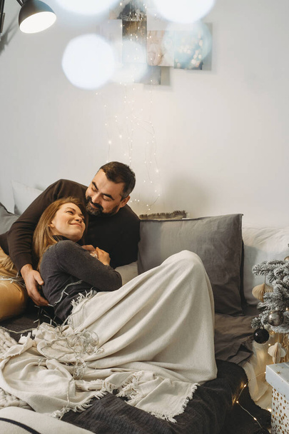 Happy couple celebrating romantic Christmas eve at home. Christmas interior decoration for family party. Copy space, greeting card. Middle-aged man and woman sitting on cozy bed with knitted blanket - Фото, изображение