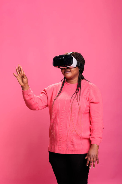 ピンクのバックグラウンドを持つスタジオで3Dエクスペリエンスを楽しむVRヘッドセットを使用してバーチャルリアリティゲームをプレイする楽しいアフリカ系アメリカ人モデル. 拡張された技術革新による電子未来的な経験 - 写真・画像