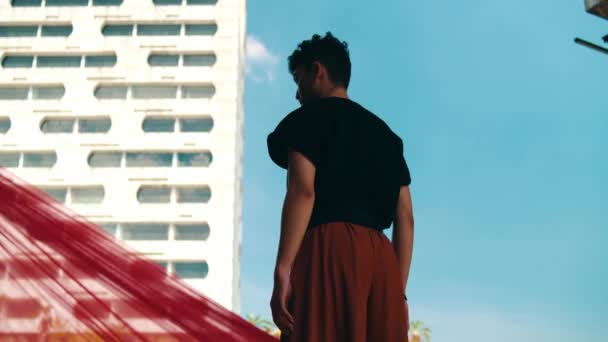 un hombre asiático de pie frente a un edificio con un cielo azul y tela roja volando durante el día - Metraje, vídeo