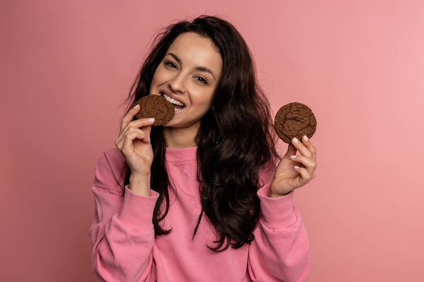 Waist-up portrait of a pleased dark-haired woman biting off a piece of oat biscuit while holding another in her hand. Sweet food concept - Photo, Image