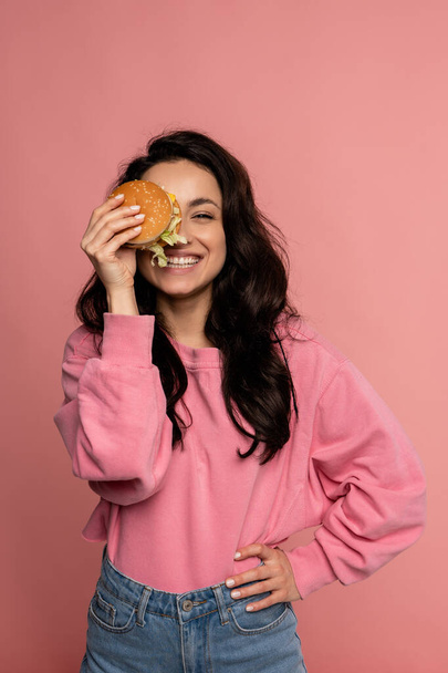 Καλά όμορφη κυρία καλύπτει το ένα μάτι με ένα νόστιμο cheeseburger, ενώ ποζάρουν στο ροζ φόντο κατά τη διάρκεια της φωτογράφισης στούντιο - Φωτογραφία, εικόνα