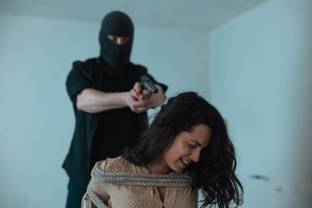 Frightened lady crying and sitting tied with a rope while kidnapper pointing a gun at her back in basement. Crime, kidnapping, violence concept - Photo, Image