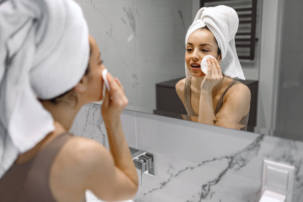 Hübsche junge Frau in Unterwäsche mit weißem Handtuch auf dem Kopf genießt eine Gesichtsbehandlung im Badezimmer und schaut in den Spiegel. Pflege, Gesundheit, Lebensstil-Konzept - Foto, Bild