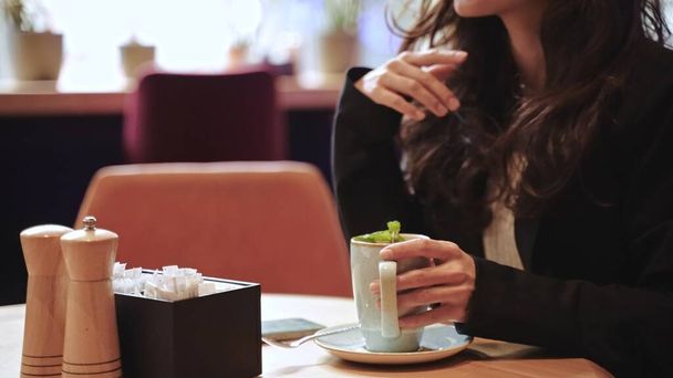 Close-up, slow motion, fingers, cup. Beautiful Caucasian girl sitting inside of modern cafe with a lot of green plants, drinking hot tea with mint with pleasure and touching her long brown hair. - Photo, Image