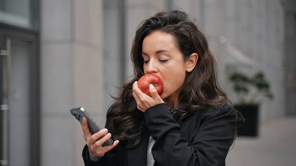 Beautiful Caucasian woman walking outdoors, wearing black jacket and white teeshirt, scrolling her smartphone and eating tasty apple. Girl walking around grey building, smiling, looking at her phone. - Photo, Image