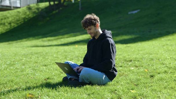 Gelukkige stijlvolle man met behulp van laptop en typen tijdens het zitten op het gazon in het stadspark. Herfstseizoen. Onderwijs, werkconcept. Langzame beweging - Foto, afbeelding