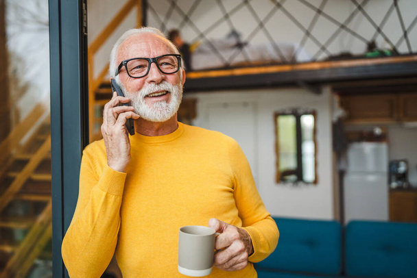 Один чоловік старший кавказький чоловік з сірою бородою стоїть на балконі біля дверей вдень щаслива посмішка використовуйте мобільний телефон, щоб зателефонувати на копію розмовного простору тримати чашку кави вранці щодня
 - Фото, зображення