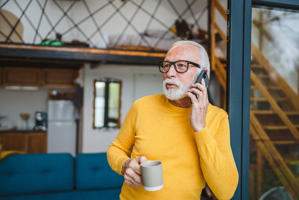 Egy férfi idősebb fehér férfi szürke szakállal áll az erkélyen az ajtóban nappal használja a mobiltelefon, hogy egy hívást talk másolás helyet tartani egy csésze kávét aggódó gondolkodó - Fotó, kép