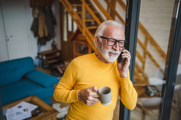 Jeden muž senior kavkazský muž s šedým vousem stojí na balkóně u dveří ve dne šťastný úsměv pomocí mobilního chytrého telefonu, aby hovor talk copy prostor držet šálek kávy ranní rutina - Fotografie, Obrázek