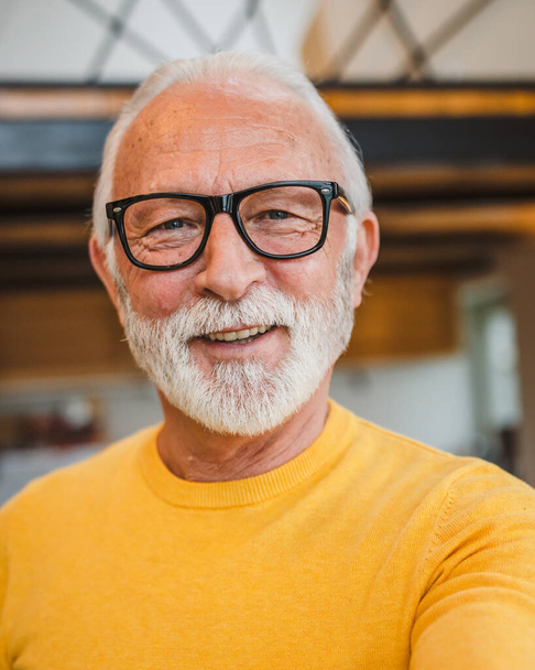 One modern senior caucasian man portrait of male grandfather with beard stand in his house in day wear yellow sweater happy confident copy space self portrait ugc user generated content - Photo, Image