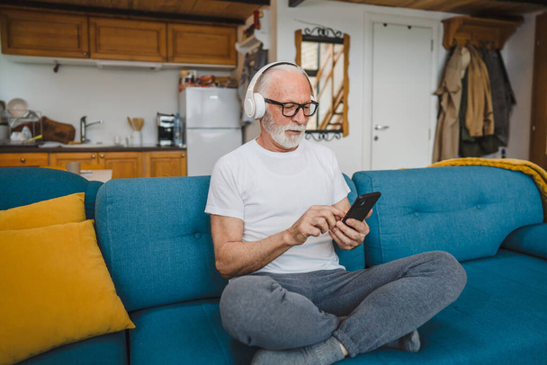 ヘッドフォンを装着した年配の男性がスマートフォンを使って自宅で音楽を演奏し - 写真・画像