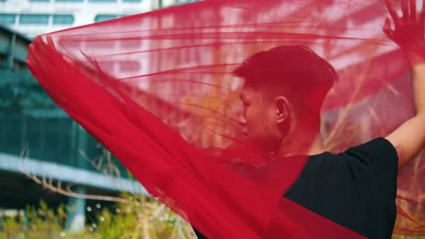 Азіатський чоловік тримає червоний шарф обома руками, коли вітер дме і здуває тканину протягом дня - Кадри, відео