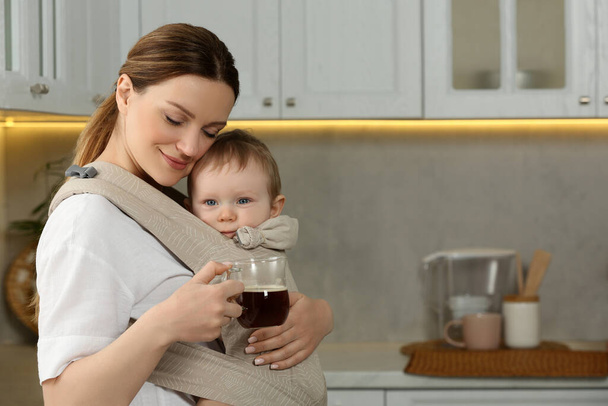 Mother with cup of drink holding her child in sling (baby carrier) in kitchen - Photo, Image