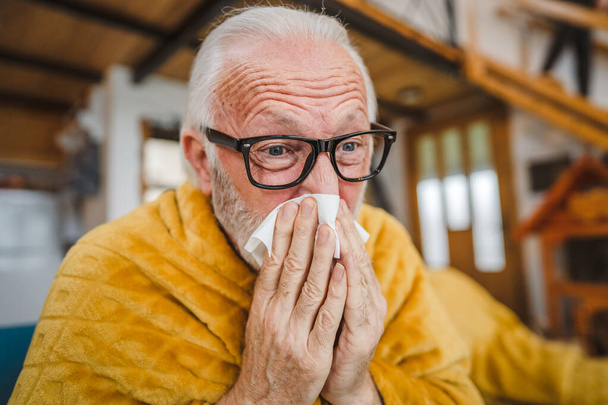 een blanke man senior man zitten op een slaapbank thuis houden Papier weefsel gemeenschappelijke koude niezen - Foto, afbeelding