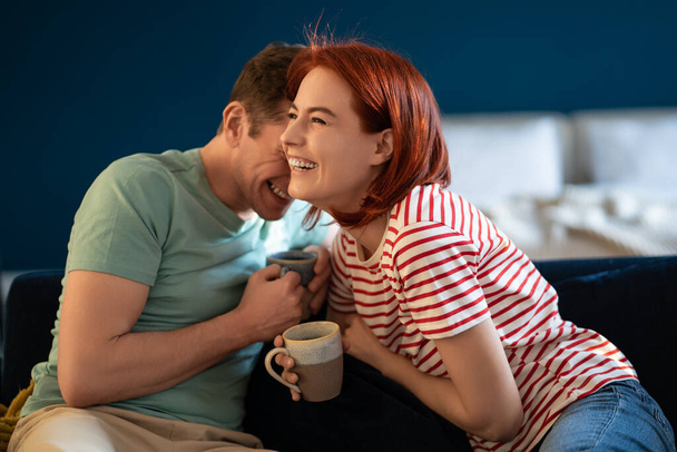 Well-rested couple talking and laughing. Middle aged smiling wife and husband sitting with cup tea morning on sofa. Loving family having fun weekend. Emotional connection in healthy relationship. - Photo, image